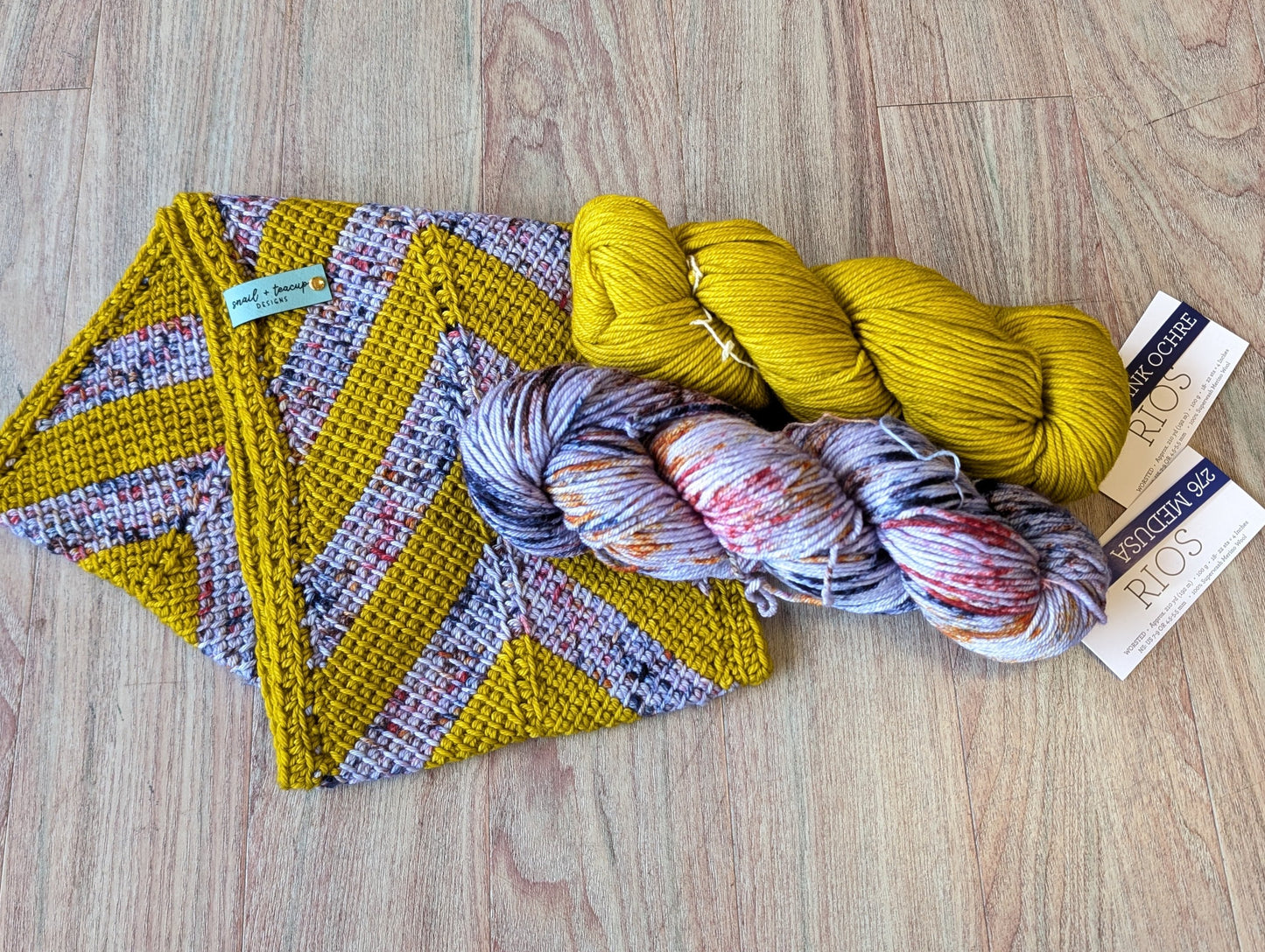 yellow and purple toned Chevron striped Cowl folded on a wooden background with two hanks of matching yarn laying on top